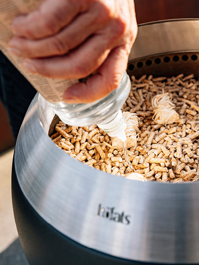 Eine Person gießt Ethanol über Holzspäne in einen Feuerkorb. Zeigt, wie man den MOON 45 anzündet.